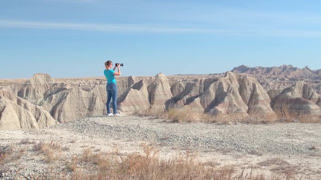 Cheerful young woman traveler taking photos of scenic Badlands National Park on sunny day in South Dakota, USA. Happy girl exploring sandstone mountain desert wilderness and photographing with camera