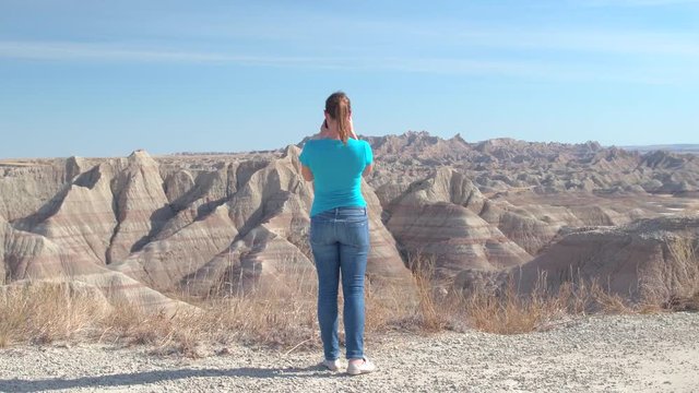 Cheerful young woman traveler taking photos of scenic Badlands National Park on sunny day in South Dakota, USA. Happy girl exploring sandstone mountain desert wilderness and photographing with camera
