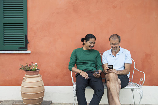Two men sitting on bench in font of orange house and looking at smartphones