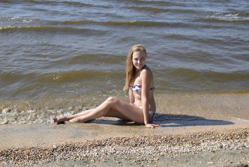 The girl the blonde in a bikini sitting on the beach in the sand. Beautiful young woman in a colorful bikini on sea background
