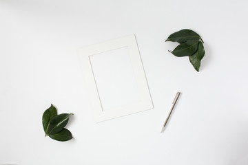 Mock up of white photo frame with leaves and silver pen on a white desktop. Flat lay.