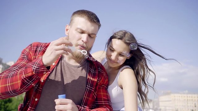 Attractive Mature man blowing soap bubbles in slow motion with his wife, the happy couple laughing sitting in the city in the sky.