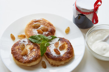 three pancakes on a white plate on white table with whipping sour cream and little jar of jam. covered with sugar powder, mint, raisins. low angle shot. isolated white on white