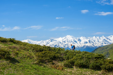 Fototapeta na wymiar Traveler ascends the green slope against the background of snow-capped mountain peaks