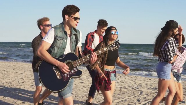 Group of young hipster friends walking and dancing together playing guitar and singing songs on a beach at the water's edge. Slowmotion shot