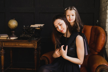 Mother and daughter hugging and playing together. Pretty little girl on beautiful woman's lap. Girls in black dresses playing in decorated room. Family weekend, beauty day, having fun, love concept.