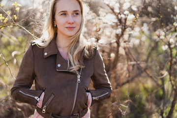 Beautiful young caucasian blonde woman in casual clothes standing near the tree in the garden. Woman portrait outdoors. Spring outfit, jeans and jacket. Copy space. Beauty, health care concept.