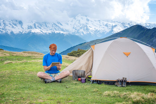 Traveler sits next to the tent, in the mountains using a tablet