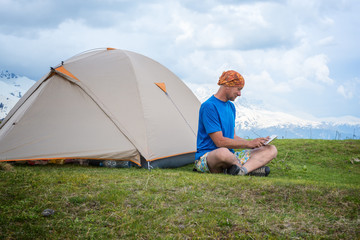 Engaged traveler sits next to the tent, in the mountains using a tablet