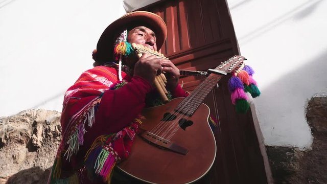Native quechua man using a colorful handcrafted chullo and a highlander hat, singing with his guitar and quena on the alleys of Cusco	