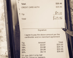 Soft focus, shallow DOF a receipt with hand written total amount and tipping from Vietnamese restaurant. Paper invoice with suggested gratuities/tips on plastic tray, open pen on top of marble table.