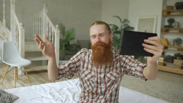 Young bearded man using tablet computer having video chat sitting in bed at home