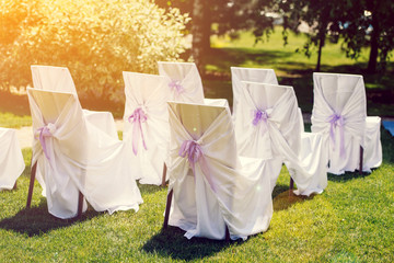 Chairs for the ceremony with white satin cape and purple ribbon on a green grass