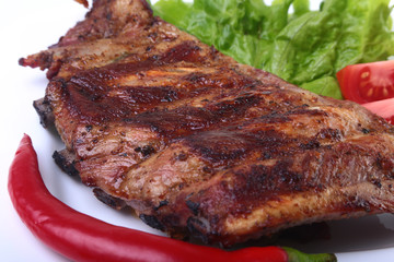 Pork ribs on grill and chili pepper with tomato, lettuce leaves on white plate