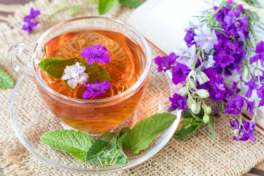 Glass cup of summer herbal tea with fresh mint and field larkspur. Bouquet of wild flowers on the notebook. Wooden table. Shallow depth of field.