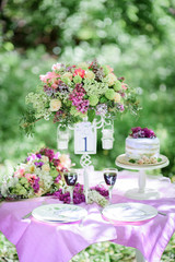 Table number one with bouquet of violet flowers and little cake on it stands in the garden