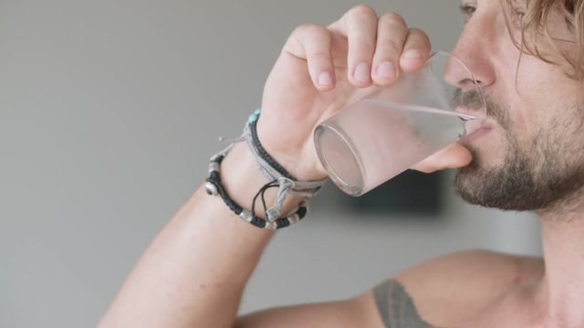 Handsome man with bare chest drinking ice water from the glass and looking out the window of the room