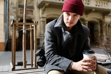 Young man in grey coat sits on bench with camera on his shoulder