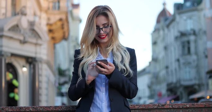 Attractive blonde woman in a business suit standing outside the office in the city center, and actively using her phone for texting, reacting happily, smiling brightly, texting back.