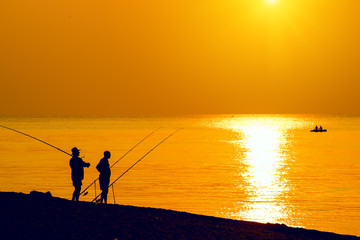 Silhouettes of fishermen with fishing rods on the beach. A boat with fishermen in the sea. Bright colorful sunset photo, dawn. Beautiful marine species.
