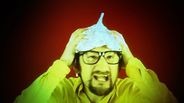 An ugly man wearing a tin foil aluminium hat (believing that it shields the brain from mind control or reading). Conspiracy, pseudoscience, paranoia. Disturbing colored shadows on his face.
