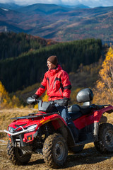 Fototapeta na wymiar Male rider in jacket and hat on a red ATV on mountain roads on a sunny day. Beautiful landscape of forest, mountains and blue sky