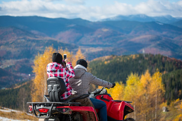 Close-up of male riding quadbike female sitting behind him makes photo of beautiful landscape mountains and forest on the phone