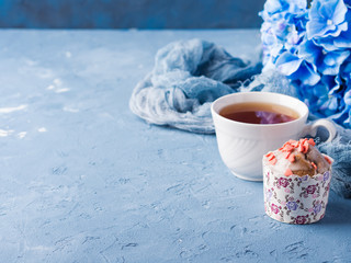 Obraz na płótnie Canvas Cup of tea on blue background with frosted cup cake, flowers and textile