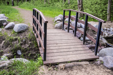 Trail leads to a small wooden bridge over the creek shore is lined with stones, in the summer forest.