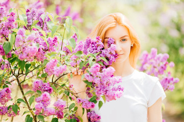 Fototapeta premium Portrait of young attractive cute redhair girl outdoor in park in blooming lilac bushes in summer sunny day. Resting among paradise flowers. Fragrance and fresh smell. Tenderness, innocence, airness.