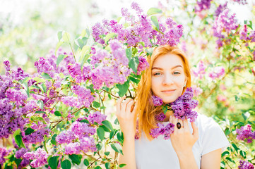 Portrait of young attractive cute redhair girl outdoor in park in blooming lilac bushes in summer sunny day. Resting among paradise flowers. Fragrance and fresh smell. Tenderness, innocence, airness.