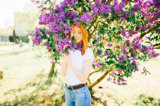 Portrait of young attractive cute redhair girl outdoor in park in blooming lilac bushes in summer sunny day. Resting among paradise flowers. Fragrance and fresh smell. Tenderness, innocence, airness.