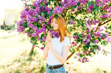 Beautiful young tender cute redhead girl with eyes closed in park in blooming lilac bushes in sunny day on weekend. Relaxing in paradise flowers. Fragrance in air. Tenderness, innocence, airness.
