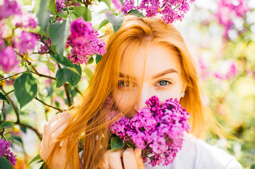 Naklejka premium Young and beautiful artistic expressive emotional face with playful and foxy, catty eyes and read hair of girl looking at camera in paradise blooming lilac bushes with flowers on nature outdoor.