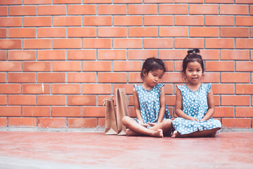 Fototapeta na wymiar Two asian kid girls sitting on the floor and waiting their parent shopping on brick wall background in vintage color tone