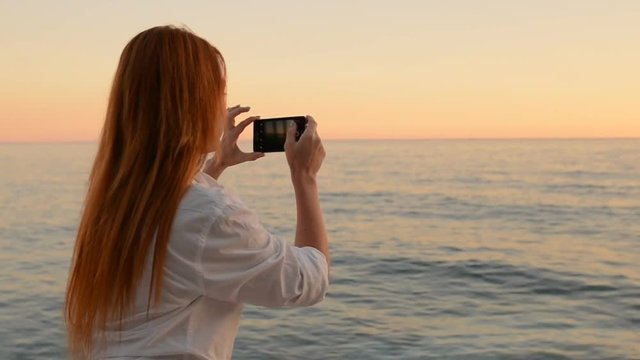 Young red-haired woman is taking pictures on the camera phone of a sea landscape by the sea, sunset