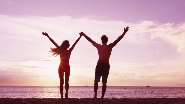 Happy couple cheering on beach enjoying sunset on summer travel vacation holiday. Happiness and bliss concept video with people raising arms of joy celebrating in silhouette.