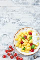 Top view of caprese gnocchi with mozzarella, tomato and basil and glass of ice water on white wooden table with empty space.