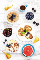 Healthy summer breakfast top view.  Oatmeal with blueberry, bowl of puffed rice cereal with pear, ricotta fritters, cup of coffee, fresh berries and grapefruit on white table.