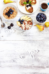 Obraz na płótnie Canvas Ricotta pancakes, bowl of porridge and puffed rice cereal with fresh berries, pear, cup of coffee on white wooden table. Top view of healthy breakfast with space for text. 