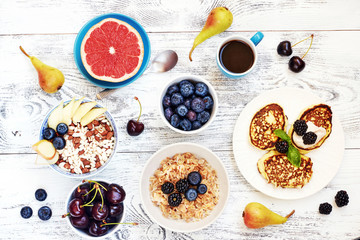 Oatmeal and puffed rice cereal with fresh berries, pancakes and grapefruit, cup of coffee and pears on white table. Top view of healthy breakfast. 