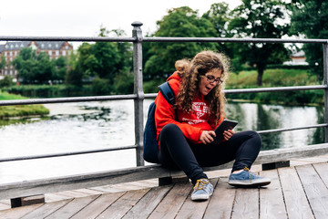 Young caucasian with  curly hair girl using tablet outdoor sitting on bridge. Student using digital tablet after school at park.