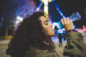 portrait young woman drinking from a bottle outdoor night life - thirsty, refresh, sipping concept