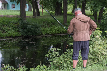  A fisherman stands on the shore of a lake pond in a forest with a fishing rod and catches fish in a jacket