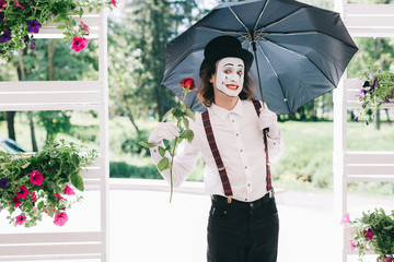 Pantomime theater artist with red rose and umbrella posing near the pot of summer flowers.