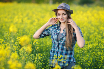 Attractive young blonde woman in blue plaid shirt straw hat enjoying her time on bright colorful blossoming yellow green meadow hard noon sun.