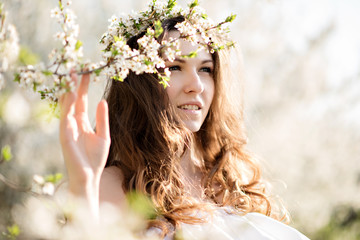 Beautiful young girl in white dress and wreath of flowers on head in blooming gardens