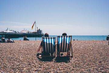 A classic British seaside scene. 2 people sat on deckchairs on stone beach