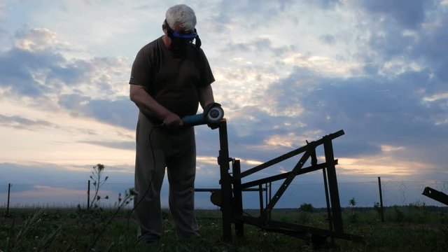 A man in age cuts off a metal angle grinder at sunset. Beautiful sky behind the worker
