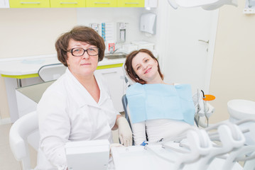 Portrait of middle aged female dentist with smiling patient sitting at dental chair, both looking at camera